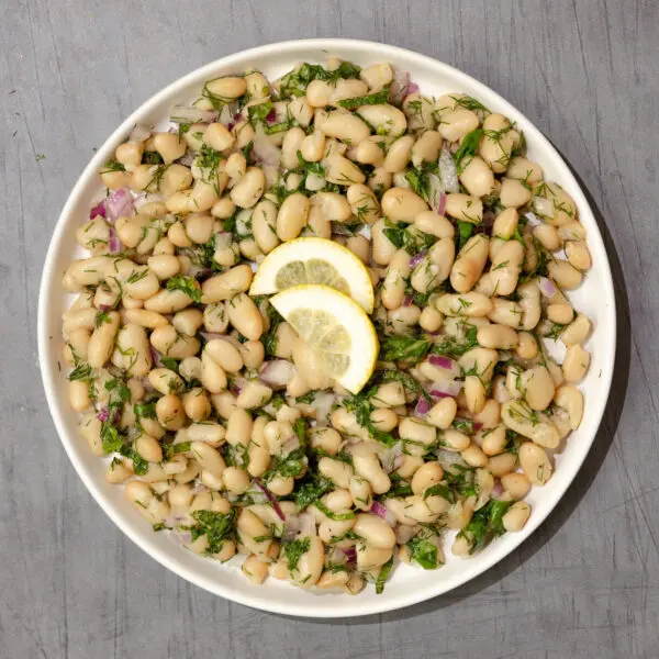 Overhead view of an Herby White Bean Salad in a white bowl garnished with two half slices of lemon and served over a gray wood table top.