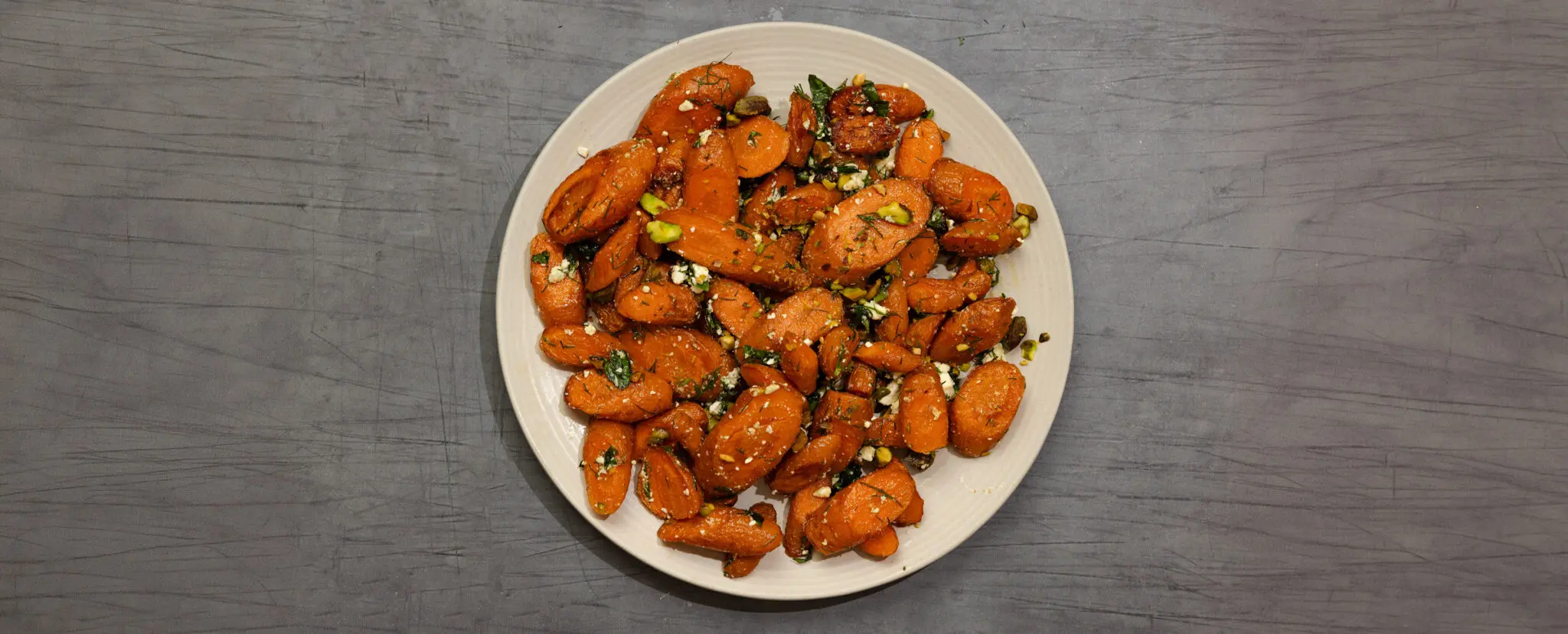 Overhead view of roasted herb feta & carrots on a round white plate, served over a gray wooden table top.