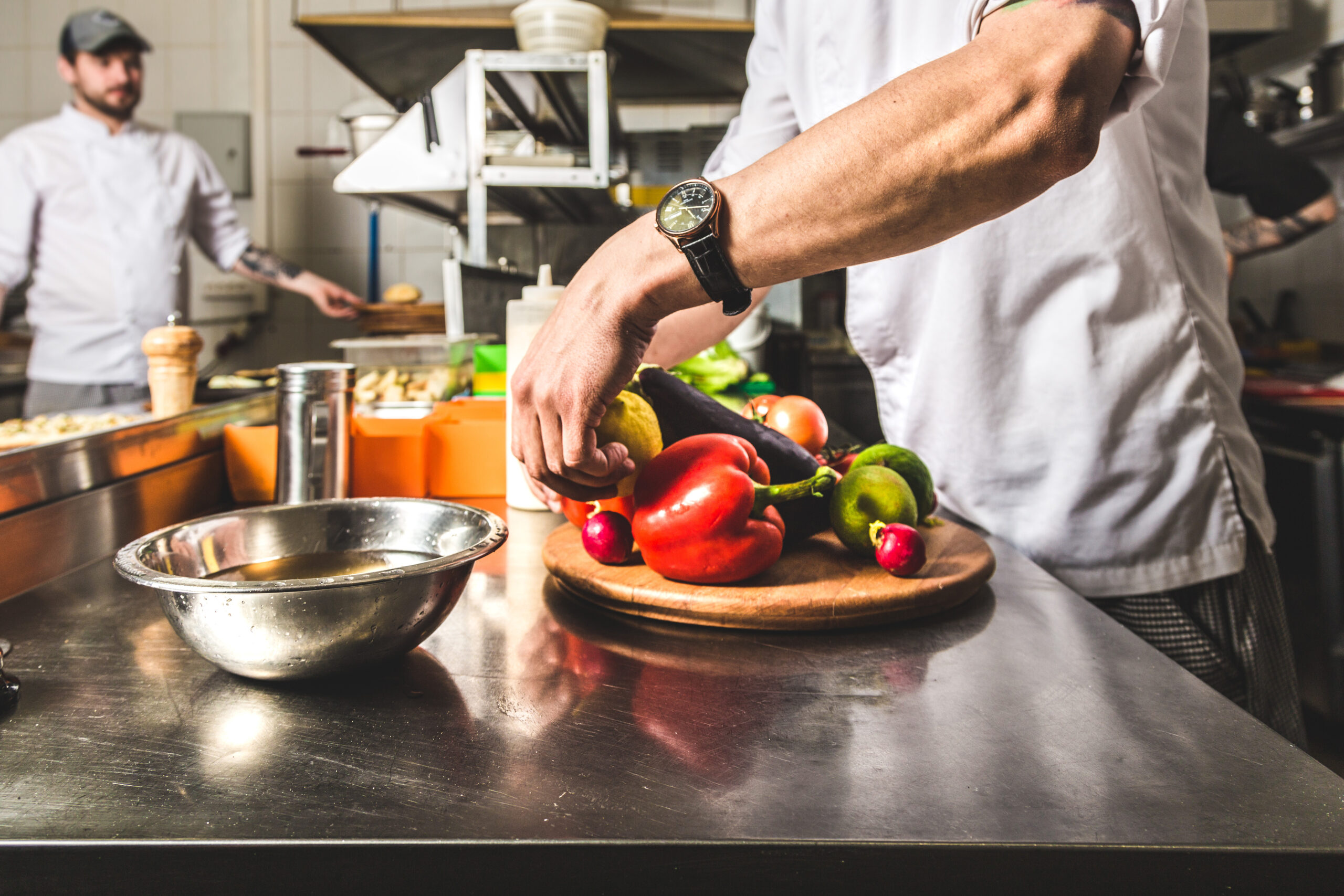 two chefs in a commercial kitchen. one is preparing fresh vegetables on a wooden chopping board.