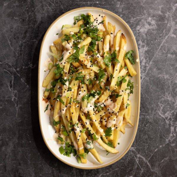 Overhead view of loaded french fries with sweet chili mayo sauce, cilantro, green onion, and sesame seeds on a large white platter, served over a black marble surface.