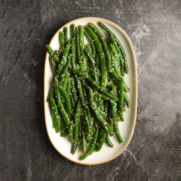Overhead view of a white platter of miso green beans served over a white gray textured stone surface.