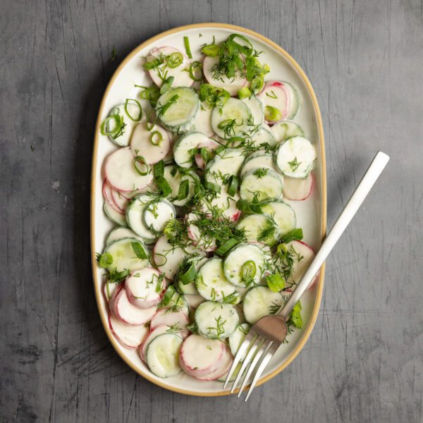 Overhead view of a large white platter of Creamy Cucumber & Radish Salad and a fork, served over a light gray textured tabletop.