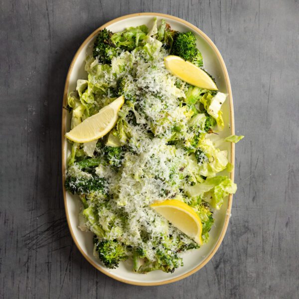Overhead view of a large ceramic platter of Roasted Broccoli Caesar Salad with lemon wedges, served over a gray textured tabletop.